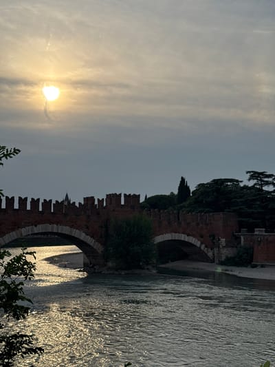 The image depicts a scenic view of the Adige River in Verona, Italy, with the Ponte Pietra bridge visible. The sun is low in the sky, casting a warm glow and creating shimmering reflections on the water, indicating it is likely sunset or sunrise. The sky is overcast with wispy clouds, but the sun is a bright, intense orb. The Ponte Pietra, a historic Roman bridge, is constructed from brick and stone, with distinctive arched openings. Lush green trees and foliage frame the scene, particularly along the riverbanks and on the bridge itself. In the background, parts of Verona's historic architecture can be glimpsed, including what appears to be a church spire. The overall atmosphere is peaceful and serene, capturing the natural beauty of the location bathed in the soft light of either dawn or dusk. No people or text are visible in the image.