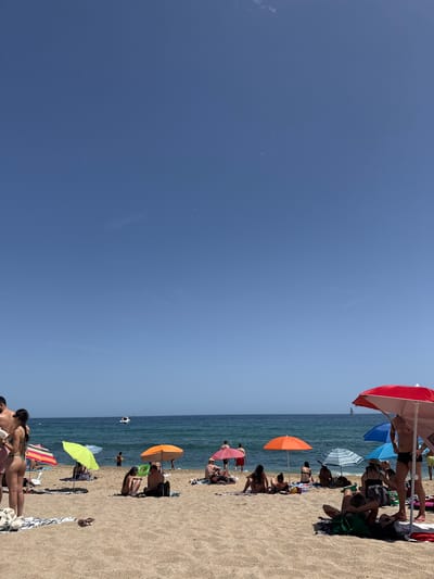 The image depicts a crowded beach in Badalona, Spain, under a clear blue sky. Numerous people are scattered across the sandy shore, enjoying the sun and the sea. Several colorful beach umbrellas, including red, orange, and green ones, provide shade. Some individuals are seated on towels or directly on the sand, while others are standing or wading in the shallow water. A few boats are visible in the distance on the calm blue sea. The beach appears to be a popular spot, with a lively atmosphere.