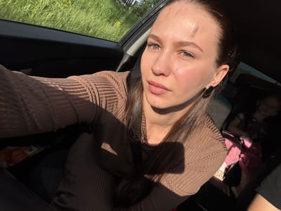 A young woman is captured in a selfie taken inside a car, likely in the front passenger seat, in Nizhnyaya Gar', Russia. She has long, dark hair, pulled back, and light green eyes. Her face is directly lit by strong sunlight, creating prominent shadows and highlighting minor marks on her forehead. She wears a brown, ribbed, long-sleeved top and a black oval earring with a gold rim on her left ear. A seatbelt strap crosses her chest.

In the background, to the left of the woman, a baby or toddler dressed in pink clothing is partially visible, possibly seated in a car seat. A leopard print pattern is visible on what appears to be a sleeve or sock near the child. The car's interior is dark, contrasting with the bright sunlight streaming in. Through the car window, a vibrant green field with scattered yellow wildflowers is visible under clear, sunny conditions, indicating daytime. There is no discernible text in the image.