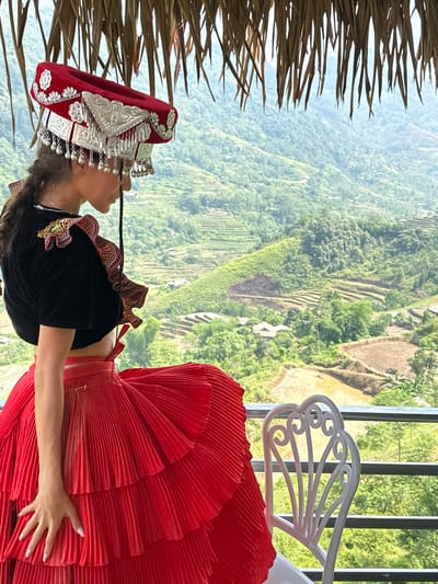A person adorned in vivid traditional attire is captured standing on an outdoor viewpoint, overlooking a majestic mountainous landscape. The subject, likely a woman, is positioned in profile, gazing out towards the horizon. She wears a distinctive large red headpiece, intricately embellished with numerous silver-colored metallic ornaments, including delicate filigree, square plates, and dangling spherical elements. Her upper garment is a black top, featuring colorful embroidered patterns on the short sleeves and across the chest, with a slight portion of her midriff visible. A spectacular, pleated red skirt, composed of multiple layers creating a tiered, fanning effect, dominates the lower half of her attire. Her left hand rests gently on the skirt, revealing light-colored manicured nails.

The background unfolds into a sprawling vista of lush green mountains and meticulously carved terraced fields, indicative of agricultural land like rice paddies. These terraces, shaped into the natural contours of the hills, are interspersed with dense vegetation. On the right side of the frame, a white, ornate metal chair and a dark metal railing are partially visible. The immediate foreground, on the left, shows dry, straw-like foliage, suggesting a thatched roof or canopy structure. The scene is brightly lit by natural daylight, implying clear weather. The image encapsulates a moment of cultural appreciation set against the breathtaking natural beauty of Thông Nguyên Commune, Vietnam. No text is visible in the image.