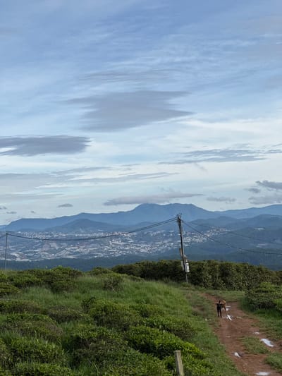The image shows a landscape in Phát Chi, Vietnam, with a view from a high vantage point. The sky is predominantly cloudy with varying shades of blue and gray. In the midground, mountain ranges are visible, fading into the distance with a hazy blue tint. A town or city sprawls in the valley between the mountains. A utility pole with wires runs along the right side of the image. The foreground shows a grassy hillside with patches of bushes and a dirt path. A person is on the path.