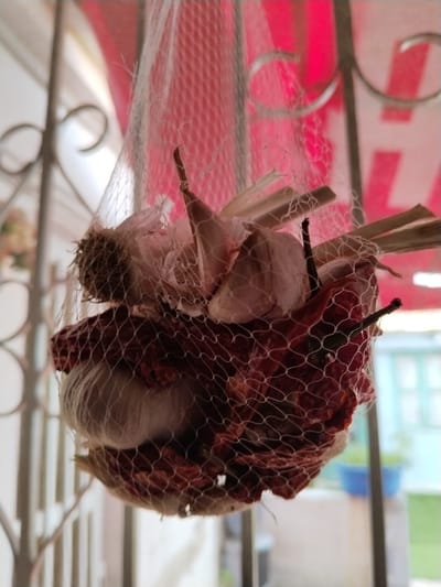 A white mesh net containing multiple light-colored garlic bulbs and numerous dark red, dried chili peppers hangs centrally. The net is suspended from a metallic, ornamental wrought-iron structure, likely a gate or balcony railing, visible as blurred, curved elements in the upper background. Behind the net, a strongly lit, out-of-focus reddish-pink fabric or awning is present. To the far right in the blurred distance, a light-colored building facade with a window featuring light blue or teal shutters or framing is visible, along with a blue potted plant. The scene takes place in the city of Sucina, Spain.