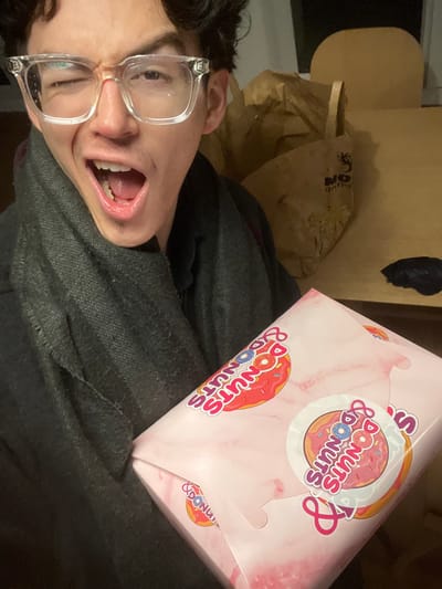 This media file captures a humorous indoor scene from Villeneuve-d'Ascq, France, featuring a person posing with a box of donuts. The main subject, a young person with dark hair and clear-framed glasses, is visible in the lower left, their face contorted in an exaggerated, wide-mouthed expression, conveying excitement or silliness. They are wearing a dark, possibly gray or black, hooded garment.
A prominent pink donut box from "Donuts & Donuts" is held in the upper right. The box features a light pink, marbled pattern and multiple logos displaying "DONUTS & DONUTS," often accompanied by a donut graphic.
In the background, behind the person and the donut box, there is a partially visible brown paper bag. Faint text, possibly "DON" or "NO ON" and a graphic resembling a spider, can be discerned on the bag. Further back, a larger, light brown object, which might be furniture or a cardboard box, is visible, along with a smaller black object.
The setting appears to be indoors, under artificial lighting, suggesting the photo was taken in the evening or at night. The individual's playful expression strongly indicates a mood of joy or anticipation, likely related to the donuts. The image itself is rotated 90 degrees clockwise.