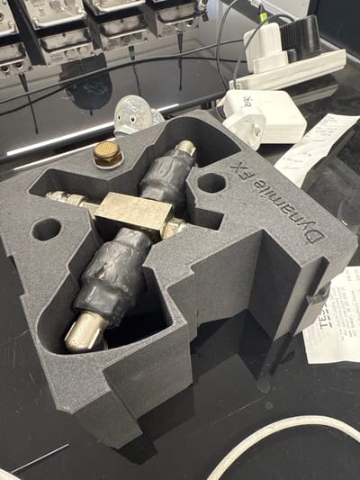 The image shows a black, foam-like object, labeled "Dynamite FX," encasing a metal fitting, possibly a valve or connector. The fitting has black coverings on either end. The foam piece is likely a custom-made container or organizer for the fitting.
In the background, various objects are visible, including electrical adapters, what appears to be a receipt from Tesco, and an array of metal components. The receipt indicates that Tesco is located in Harlow Church Langley Superstore. Given that Harlow is a town within the Epping Forest district of Essex, United Kingdom, the image could be taken in this general locale.
