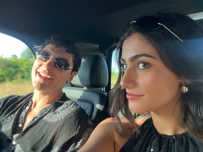 This is a close-up, slightly overexposed selfie of two people, a woman and a man, in a car. The image is rotated 90 degrees counter-clockwise. The woman is on the left, looking towards the camera with a slight smile. She has dark hair, visible earrings, and is wearing a black top with sequins. The man is on the right, also looking towards the camera with a wide smile, showing his teeth. He has dark, curly hair, is wearing sunglasses and a black shirt with a white pattern. They are seated in what appears to be a car, with car seats visible in the background. The scene suggests a sunny day, possibly late afternoon given the warm lighting. The background shows a blurred view of greenery and sky, suggesting they are traveling through a scenic area, possibly in Santa Eulària des Riu, Spain. There are no visible text elements in the image.