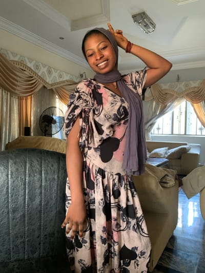 The image shows a smiling woman posing indoors, likely in a living room setting. The woman, who has dark skin, is positioned slightly to the left of the frame, facing the viewer with a broad, cheerful smile that shows her teeth. She is wearing a light-colored dress with a distinctive pattern featuring black hearts, pink accents, and black or grey floral motifs. A solid grey or light purple head covering is wrapped around her head and neck. On her right wrist, she wears several thin, reddish-brown bracelets. Her right arm is raised, with her hand forming a peace sign (V-shape with index and middle fingers), while her left hand rests casually near her waist with fingers slightly curled.
The background reveals a home interior. To the left, a silver or metallic rectangular light fixture with a slatted design is visible on the ceiling. Light-colored walls, possibly white or off-white, frame the scene. Behind the woman, decorative light-colored curtains, possibly cream or beige, with valances and drapes, are visible, suggesting a window letting in natural daylight. A black standing fan is clearly visible in the mid-ground to the right of the woman. In the foreground on the right, a dark-colored, possibly leather or faux-leather, textured armchair or sofa is prominent. Further in the background, several lighter-colored upholstered sofas or armchairs are discernible, some appearing to be recliners. The overall impression is one of a relaxed and happy moment captured in a brightly lit indoor space during the daytime.