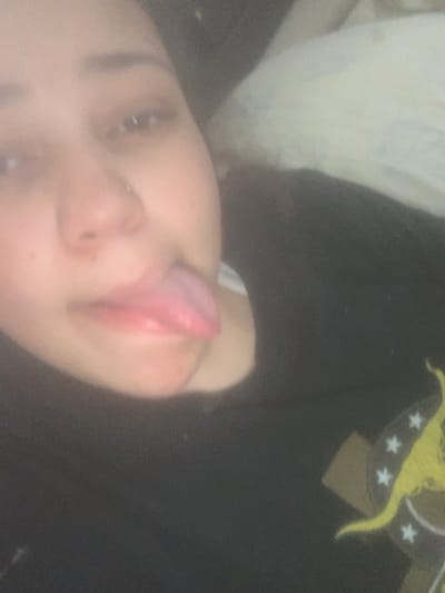 A blurry, dimly lit indoor photograph depicts a female individual from the chest up, captured in the city of Lancaster, United States of America. She has a light to medium complexion and visible dark hair. A silver-colored stud piercing is present on her left nostril. Her mouth is open, and her pink tongue is extended outwards. She wears a dark-colored garment, likely a t-shirt, under which a light-colored strap is visible near her chin. A yellow and white graphic design, possibly depicting an animal (resembling a kangaroo) surrounded by stars, is partially visible on the lower right portion of her attire. The background is indistinct and appears to contain light-colored fabric or bedding on the upper right, consistent with an interior personal space.