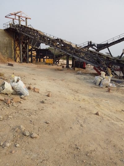 This image depicts an aggregate processing facility located in Hoche, Nigeria. The scene is dominated by a large, multi-tiered industrial structure featuring inclined conveyor belts, supported by dark metal framework and trusses, and a concrete retaining wall on the left. A yellow generator or power unit with "PELIC" visible on its side is situated beneath the conveyor system in the midground. The ground consists of dry, light-brown earth, uneven and scattered with small rocks and debris, including pieces of red brick. In the left foreground, two partially filled white sacks, one marked "BENGAL CEMENT," lie on the ground. In the right foreground, a stack of approximately six to seven additional white sacks is present. No people are visible. The background shows sparse vegetation under an overcast or hazy sky.