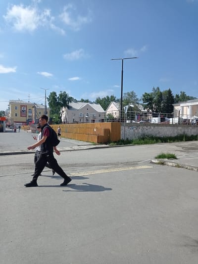 This is a full, eye-level, outdoor, medium-shot photograph taken on a sunny day in the city of Degtyarsk, Russia. The scene depicts a street with a pedestrian crossing. In the foreground, two young men are walking across the street from right to left. The man on the right is wearing a black vest over a burgundy shirt and black pants, carrying a black backpack. The man on the left, partially obscured, is wearing a white shirt and dark pants. They are walking on a paved road with a yellow tactile paving strip indicating a pedestrian crossing. In the midground, there is a low, orange wooden fence separating the street from a raised area with grass and a concrete wall. Beyond the fence, several buildings are visible, including a light-yellow building with a portrait of a man on its facade, and other residential buildings with light-colored walls and white roofs. Trees are visible behind the buildings. A street light with two lamps stands tall in the background. The sky is clear blue with a few scattered white clouds.