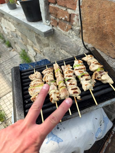 The image captures an outdoor grilling scene in Krumovo, Bulgaria. Six wooden skewers, laden with cubed chicken, onion pieces, and green bell pepper segments, are arranged on a black metal grill grate. The chicken displays visible char marks, indicating it is in the process of being cooked. A light-skinned human hand is positioned in the lower-left foreground, with the index and middle fingers extended in a V-shape, partially obscuring the grill. The background on the left consists of a low stone wall with a concrete cap, beyond which a tiled ground with sparse vegetation is visible, along with a partially obscured blue plastic crate. On the right, a reddish-brown brick wall and a darker, textured stone surface are present, intersected by a black electrical cable. A white cloth with light blue patterns is visible beneath the grill, serving as a covering for the surface below. Two out-of-focus potted plants are situated on the stone wall in the upper-left background.