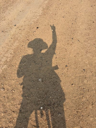 A shadow of a person wearing a cowboy hat stands on a dirt ground. The person's right arm is raised with the index finger pointing upwards. The shadow is cast on a surface composed of sand and small pebbles, with some larger stones scattered throughout. The lighting suggests it is daytime, likely with the sun positioned behind the person. There are no other visible people or objects in the immediate vicinity of the shadow. The overall impression is one of an outdoor, possibly rural or undeveloped area, due to the natural ground composition. No specific location in Myandigi, Russia can be determined from the visual information provided in the image.
