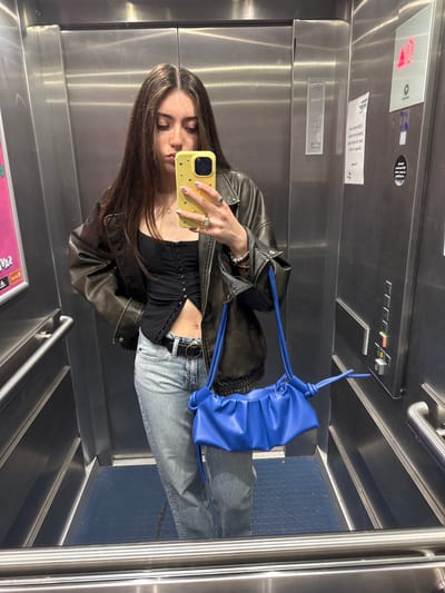 A young woman is depicted taking a mirror selfie inside an elevator. She stands in the center of the frame, holding a bright yellow smartphone with a patterned case to her face, partially obscuring her features as she looks into the phone's screen. She has long, dark hair with lighter highlights.

She is dressed in a dark brown or black leather jacket, a black ruched crop top exposing her midriff, and light blue ripped jeans with a dark belt. A vibrant royal blue, gathered-style handbag with a distinctive rope-like strap is slung over her left arm. She wears rings on her right hand, including a prominent gold-toned one, and bracelets on her left wrist. Her fingernails have white tips adorned with small black dots.

The elevator interior features reflective stainless steel walls, typical of modern lifts, which reflect the subject. The floor is covered with a dark blue, textured mat featuring circular raised patterns, and chrome handrails are visible on the sides. Above her right shoulder, an elevator control panel is visible with black buttons indicating floor numbers (1 through 6, and additional function buttons like a bell and an orange-colored one). A digital display above the buttons shows a red "1", indicating the current floor. Several white rectangular stickers with mirrored, largely illegible text, and a circular black sticker are affixed to the steel wall, including one that partially shows "E1216". The setting is an indoor location, specifically an elevator in Hull, United Kingdom.