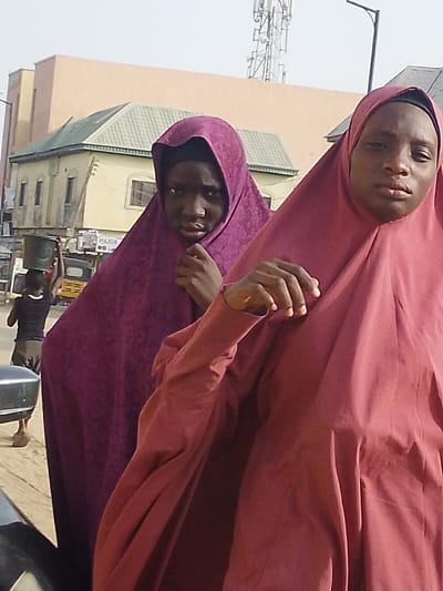 The image depicts a daytime street scene in Garun Gawa, Nigeria. In the immediate foreground, two individuals, likely female, are centrally positioned. The individual on the right wears a reddish-brown full-length garment and matching hijab, looking directly at the viewer with a neutral expression. Her right hand is visible near her upper chest. To her left and slightly behind, a second individual wears a purple full-length garment and matching hijab, also looking towards the viewer with a focused expression; her hands are near her chest. In the left background, a person with their back partially turned is seen walking away, carrying a dark bucket on their head. A yellow motorized tricycle is visible further back. Buildings line the background, including a multi-story structure with a green-tiled roof and a larger, peach-colored building. A tall metal telecommunication tower and a street light pole are visible against a clear sky in the upper background. A dark-colored vehicle's partial side is visible in the bottom left foreground.