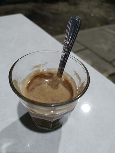 The image shows a close-up of a glass of coffee with a spoon in it, placed on a white table. The coffee has a frothy top and is a rich brown color. The glass is clear and hexagonal in shape. The spoon is made of metal and has an ornate design on its handle. The background is dark and appears to be outdoors at night, with some blurred details of pavement and foliage. There is a bright light source reflecting on the table, possibly from a lamp. The overall scene suggests a moment of quiet enjoyment, perhaps late at night. The location is Keudepanteubreuh, Indonesia. No text is visible in the image.