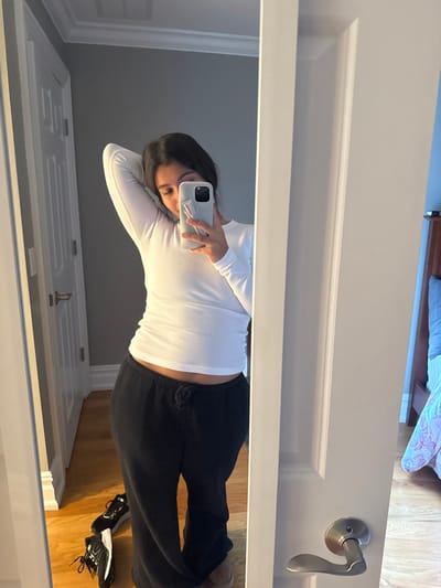 The image displays a female individual taking a mirror selfie indoors, located in a residence within Princeton, United States. The individual, possessing dark hair, is reflected from approximately the mid-thigh upward. She is wearing a white, long-sleeved shirt that exposes a portion of her abdomen, and black sweatpants with a visible drawstring. Her right hand holds a light-colored smartphone, equipped with a white grip accessory, positioned to obscure the lower part of her face. Her left arm is raised with the hand resting behind her head.
The reflection reveals a room with medium-gray walls, white crown molding, and a white paneled door featuring a silver lever handle, visible on the left side. The flooring throughout the visible areas is light-colored hardwood. Near the individual's feet, two dark-colored athletic shoes are present on the floor, one oriented on its side. To the right of the camera's perspective, through an open white door frame, another room is partially visible. This room features light blue walls, a dark brown wooden headboard, a blue pillow, and a multi-colored patterned comforter on a bed. A silver lever door handle is also visible on the white door frame in the foreground, separating the space where the photograph is being taken from the room with the bed.