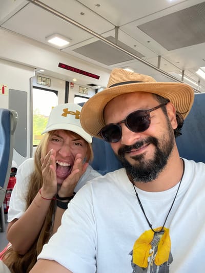 The image is a selfie taken from a low angle inside a train. Two people, a man and a woman, are the subjects. The man is on the left, wearing a straw fedora hat and sunglasses, with a beard. He is looking towards the camera. The woman is on the right, wearing a white baseball cap with a gold logo and is making a surprised or excited expression, with her hands near her face. Both are wearing white t-shirts. The man has a black cord necklace with a silver tag that says "RAW". The woman has a red bracelet on her wrist.
The interior of the train is visible, with light-colored walls and blue seating. There are overhead lights and a red digital display on the wall, which appears to show "HI HII 1 2 37". There are also safety signs on the wall. Outside the train windows, greenery and some buildings can be seen, suggesting a daytime setting, likely sunny. The overall emotion conveyed is one of fun and excitement, possibly during a trip. The provided location context suggests the train is in or near Sant Adrià de Besòs, Spain.