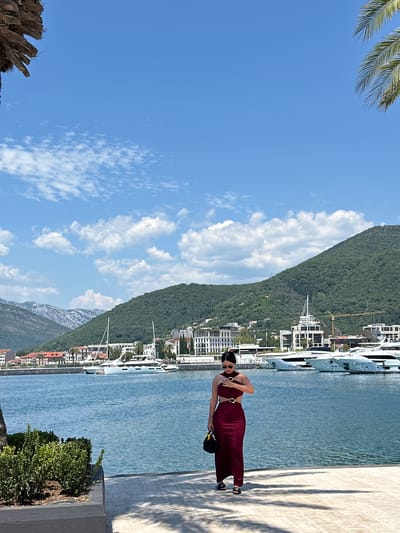 A woman wearing a dark red dress with cutouts and gold buckles walks along a waterfront promenade. She holds a black bag in her left hand and touches her chest with her right hand. She wears black sandals and sunglasses. The promenade is paved with light-colored tiles, casting shadows from nearby trees onto the ground.
In the background, a calm body of blue water is filled with docked yachts and sailboats of various sizes. Beyond the water, a city skyline features white buildings, some with red roofs, nestled against the base of lush green, tree-covered mountains. The sky is a bright blue with scattered white clouds. The location is identified as Donja Lastva, Montenegro. Part of a palm tree trunk is visible on the left side of the image, and palm fronds are visible in the top right corner.