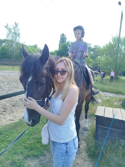 In Novohuivyns'ke, Ukraine, a young girl in a blue riding helmet sits atop a brown horse, holding the reins. Beside the horse, a woman with blonde hair and sunglasses smiles at the camera, her hand resting on the horse's muzzle. She wears a white eyelet top and light blue jeans. The scene appears to be a rural or equestrian setting, with a sandy arena, grass, and trees in the background. Two other figures are visible further back, one on horseback and another standing. A wooden structure and fence are also present.