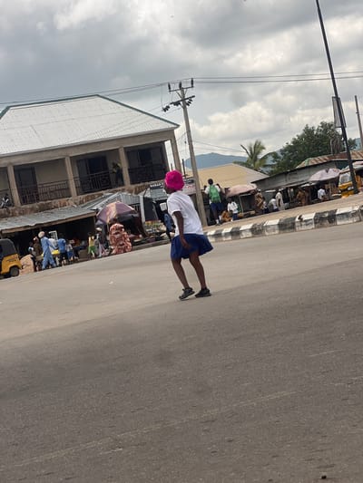 This is a vertical, slightly tilted, eye-level shot of a street scene in Gulum, Nigeria, under a cloudy sky. The foreground is dominated by a paved road, with a person in the middle ground, facing away from the camera. They are wearing a white shirt, blue shorts, and a bright pink hat, with black shoes. To the left of the road is a building with multiple stories and shops on the ground floor. These shops appear to be open, with goods displayed. There are several other people in the background, some interacting with vendors, others walking. Some umbrellas are visible, suggesting a market or outdoor shopping area. A yellow utility vehicle, labeled "PT-4011", is parked to the far left. Visible text includes "PT-4011", "BABAN F", "CASH DEPOSIT", "DATA SUBSCRIPTION", "AIR-TIME RECHARGE", "ICATIO", "SUG", "SERVICES", and "ICE FISH". The scene suggests a typical day in a busy commercial area. The time of day is indeterminate due to the overcast conditions.
