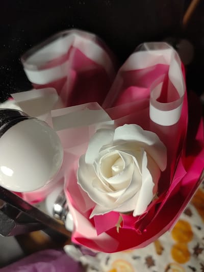 This is a close-up, overhead shot of a single white artificial rose. The rose is displayed in a wrapping of pink and white material, creating a bouquet-like presentation. The background is dark and out of focus. On the left side of the image, a glossy white sphere, possibly a decorative element, is partially visible. The overall impression is of a gift or decorative item. The image does not provide any information about the city of Saransk, Russia, or its location within the city.