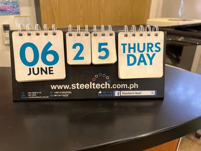 This media file is a close-up photograph of a desk calendar placed on a dark, reflective surface, likely a tabletop. The calendar displays the date "06 JUNE" on the left, followed by "25" and then "THURSDAY" on the right. The numbers and text are in a bold, blue font. Below the calendar, a website address "www.steeltech.com.ph" is prominently displayed in white text. Further details include contact information: "Tel : 567.3668 to 79", "Fax : 567.3503", and "Email : info@steeltech.com.ph". There are also other contact details like "1.800.10.ROOFING" and a mobile number. A Facebook integration is visible with "Like us on Facebook" and a Facebook logo next to a search bar labeled "Steeltech Roof". The background shows some out-of-focus office equipment, including what appears to be a white appliance and dark cabinetry. The lighting suggests an indoor setting, possibly during daytime. The overall scene appears to be a workspace or reception area, given the presence of office-related items and contact information for a business, Steeltech Roofing. The image's resolution is clear, allowing for easy reading of the text.