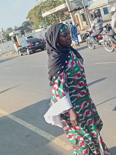 An older woman wearing a green, red, and white patterned dress and black headscarf stands on a sunlit street. A white shirt is visible under the dress's sleeves. Cars and motorcycles populate the background, along with buildings and trees. A sign for "MUHARN" is also visible. The woman appears to be looking upwards. The scene is captured from a slightly elevated angle.