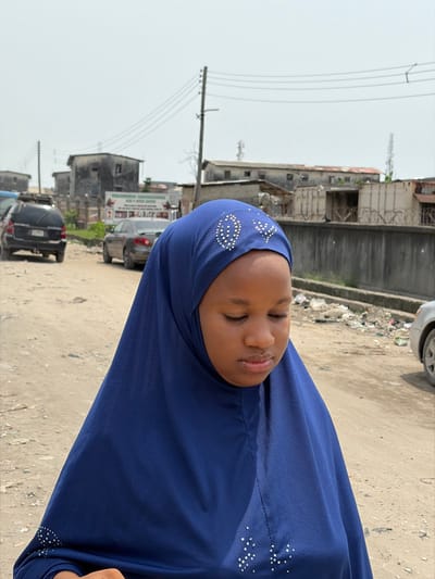 A young individual, likely female, stands on an unpaved dirt road in Jakande, Nigeria, under a bright, hazy sky. She wears a deep blue hijab, which covers her head and drapes over her shoulders and chest, adorned with silver or white rhinestone patterns including an oval-like design and a heart-like shape on the forehead area, and scattered dots on the lower front. Her eyes are closed or cast downwards, and a subtle nose piercing is visible on her left nostril.
The immediate foreground consists of light brown, dusty ground. In the background, an unpaved road extends, with several vehicles parked along its left side, including a dark SUV and a light-colored sedan. Further right, a partial view of a light-colored car's front bumper is visible. A large, faded billboard or sign with text and images stands between the vehicles and distant structures. Multiple utility poles with numerous overhead power lines traverse the scene horizontally. Along the right side of the road, a concrete wall topped with barbed wire separates the road from accumulated trash and debris. Behind this wall, and further in the distance, are several dilapidated, multi-story buildings under the hazy sky.
