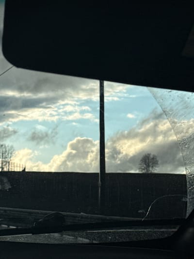 The image is a landscape-oriented shot taken from inside a vehicle, looking out through a side window. The left side of the frame is dominated by a dark, featureless interior component of the vehicle, possibly a pillar or a door panel.
The central part of the image shows a rectangular window, which is visibly dirty with streaks and dust, particularly in the upper section. A prominent dark, horizontal bar divides the window, suggesting a frame element or part of the vehicle's structure.
Through the window, a vibrant sky filled with white and light blue clouds is visible. Many clouds have golden edges, indicating the presence of low sunlight, suggestive of either sunrise or sunset. Bare tree branches are faintly silhouetted against the sky, visible in the upper right and lower right portions of the window's view.
On the far right of the image, beyond the window, dark exterior elements are present. These include a blocky, dark structure resembling a building or wall, a partially visible fence or railing at the bottom, and a dark, curved component that could be a part of the vehicle's exterior, such as a mirror housing or a wiper mechanism for an adjacent window. A thin, light-colored line, possibly a wire, is also visible against the dark exterior. No people or text are visible. The weather appears clear with significant cloud cover, and the bare trees suggest a season like autumn or winter. The location context is Aleksandrovskoe, Russia.