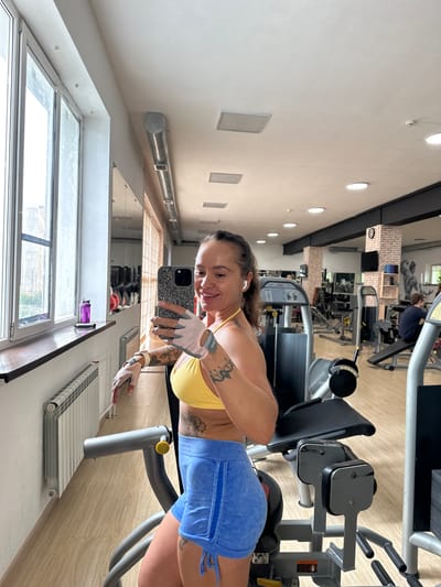 A woman is taking a selfie in a gym. She is wearing a yellow sports bra and blue athletic shorts, with fingerless gloves on her hands. She has tattoos on her arms and back. She is smiling at the camera. The gym appears to be well-equipped with various weight machines, and there is a large window in the background. The lighting suggests it could be daytime. The overall mood seems positive and energetic.