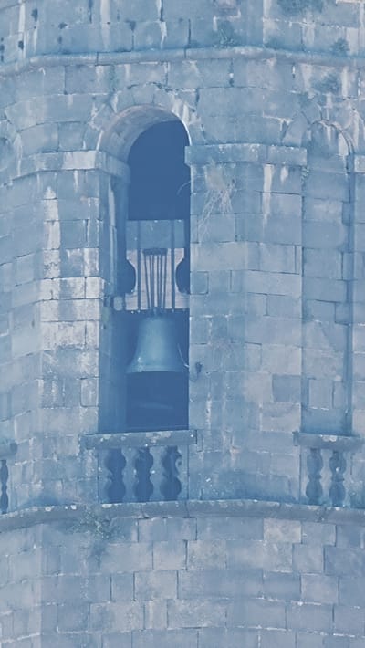 The image shows a close-up view of a bell tower in the town of Rupit i Pruit, Spain. The tower is constructed of large, rough-hewn stone blocks, giving it a rustic and ancient appearance. In the center of the image, a large bronze bell is visible, hanging within an arched opening of the tower. The bell appears old and weathered, with a patina that suggests age. Above the bell, there is a wooden structure that likely supports it, with vertical wooden elements. Below the bell, there is a small balcony with decorative stone balustrades. The overall scene is characterized by the stone architecture and the prominent bell, conveying a sense of historical significance and a connection to the past.