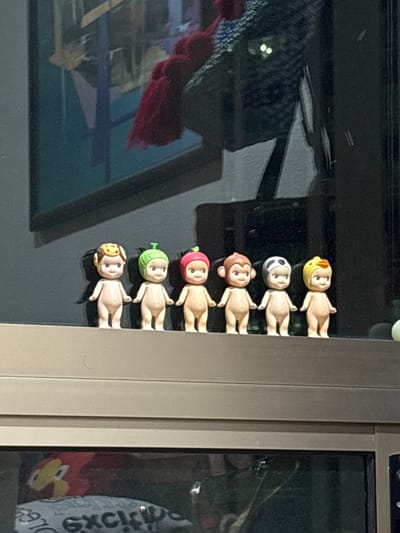 The image shows a row of six small, humanoid figurines with different colored head coverings attached to the frame of a window. The figurines are pale with simple facial features and no clothing. From top to bottom, the head coverings are yellow, black and white, brown, red, green, and tan. The window frame is gray. Reflections are visible in the window glass including a blurred image of what appears to be a red and blue object with tassels. Part of a white bag with black writing is visible in the adjacent window. The location is Jesús del Monte, Mexico.