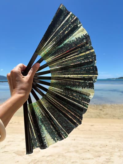 A close-up, eye-level shot shows a person's hand holding a partially opened black-ribbed fan with a dark, textured pattern on the fabric. The fan is held against a bright blue sky, with the ocean and a sandy beach in the background. The fan's pattern appears to be an abstract representation of trees or foliage, with streaks of dark green, yellow, and white. The hand is positioned to the left of the frame, with the thumb gripping the fan's central spine and fingers spread to support the fabric. The person is wearing a light-colored bracelet on their wrist. The beach is light brown and appears dry, with some scattered shadows. The ocean is calm and a lighter blue, with a faint horizon line visible in the distance. The scene is illuminated by bright daylight, creating a high contrast between the fan and the background. The location is identified as Baan Thong Krut, Thailand.