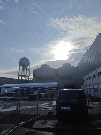 The image depicts an industrial area with large, modern buildings. In the foreground, a dark-colored van is parked, its rear visible, with the license plate NW-1961. A temporary fence with metal posts and mesh panels surrounds an area with puddles on the ground. Behind the fence, stacks of materials, possibly construction-related, are visible. In the mid-ground, a large, spherical water tower stands on a metal structure, with industrial buildings behind it. The sky is partly cloudy, with the sun partially obscured by clouds. The scene appears to be in the city of Charleroi, Belgium, based on the license plate.