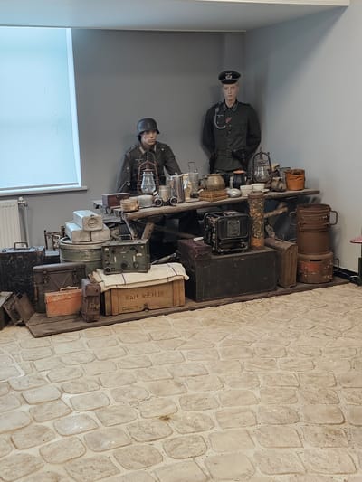 The image shows a museum display, likely in Архангельское, Russia, featuring two mannequins dressed in what appears to be World War II-era German military uniforms. One mannequin wears a helmet, while the other has a peaked military cap. They stand beside a shelf laden with various pieces of vintage military equipment. These include lanterns, metal canisters, wooden boxes, and communication devices such as radios. The backdrop is a plain gray wall on one side and a faux-stone wall on the other. A window is visible to the left.