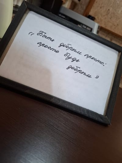 A framed handwritten quote is placed on a dark brown wooden surface. The quote, written in black cursive, reads "Быть добрым просто: просто будь добрым". The frame is dark gray and made of wood, with a visible grain pattern. The background shows a blurred out room with wooden paneling and some equipment. The overall scene suggests an indoor setting, possibly a cafe or office space. No people are present.