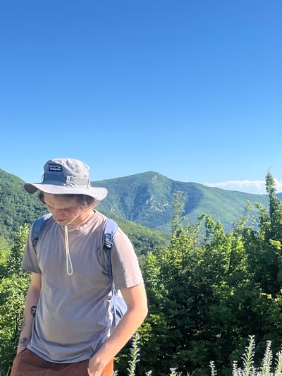 The image shows a young person wearing a grey bucket hat with a Patagonia logo and a light brown t-shirt, standing in a mountainous, forested area under a clear blue sky. They are wearing a blue backpack and brown shorts. The person is looking down, with their right hand in their pocket. The background features lush green trees and rolling hills, characteristic of the landscape found in Petrovac-na-Mlavi Municipality, Serbia.