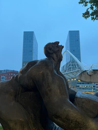 The image shows a low-angle view of a large, dark bronze sculpture in the foreground, depicting a human figure with its head tilted back and mouth open in a scream or cry. The sculpture is positioned in front of a modern cityscape. In the background, two identical, sleek glass skyscrapers stand tall against a pale blue, overcast sky. Between the skyscrapers, a contemporary white cable-stayed bridge with a distinctive arching structure spans across a body of water. Below the bridge, a building with the illuminated sign "dermitek" is visible. The scene appears to be set in Bilbao, Spain, likely near the river. The overall atmosphere is somber, possibly due to the lighting conditions and the expressive sculpture.