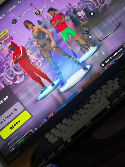 The image shows a computer monitor displaying a video game lobby. Three characters are visible on the screen, two human characters and a dog. One human character is a woman with brown hair, wearing a grey tank top and brown pants. Another human character is a man with dark skin, wearing a red and green outfit. The dog is grey and appears to be a pit bull. The characters are standing on what looks like hoverboards or skateboards. In the background, there is a city-like environment with buildings and a Ferris wheel.

On the left side of the screen, there are user interface elements, including player names like "Mukata XD" and "Nakata-G14". A prominent yellow button with the word "READY" is visible. Other UI elements include "Build, Squad, Fill" and "60 FPS".

The monitor itself is partially visible, with a keyboard in the foreground. The scene is illuminated by the light from the monitor.

The location of Kukorevo, Bulgaria is not depicted or suggested in this image.