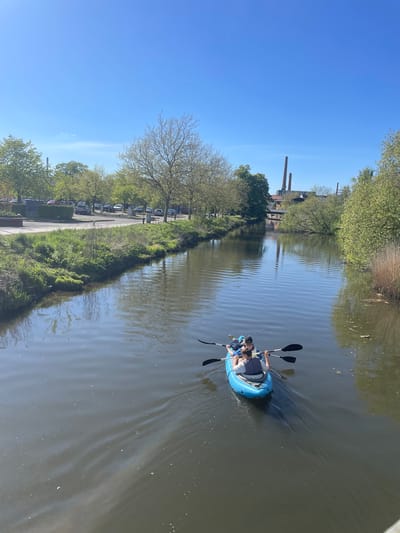 A horizontal shot shows two people in a blue inflatable kayak on a waterway. The waterway reflects trees and two brick chimney structures on the left side. The kayak occupants are paddling, creating ripples in the water. On the left bank, a path runs parallel to the water, lined with grass, bushes, and parked cars. The sky is blue. The two brick chimney structures are located at the former industrial complex of Kolding Sydbanegade.
