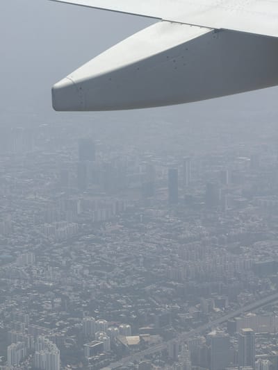 An aerial view shows a dense urban landscape, likely Gurgaon, India, shrouded in haze or smog. The foreground is dominated by the wing of an aircraft, a large, white, aerodynamic structure. Below, a sprawling city unfolds, characterized by numerous buildings of varying heights and architectural styles. Many structures appear to be apartment complexes or office buildings, forming a dense grid of urban development. Roads and infrastructure, including what seems to be a highway or elevated road, are visible weaving through the city. The overall impression is one of significant urban expansion and a high population density. The atmospheric conditions obscure distant details, giving the scene a muted, gray appearance.