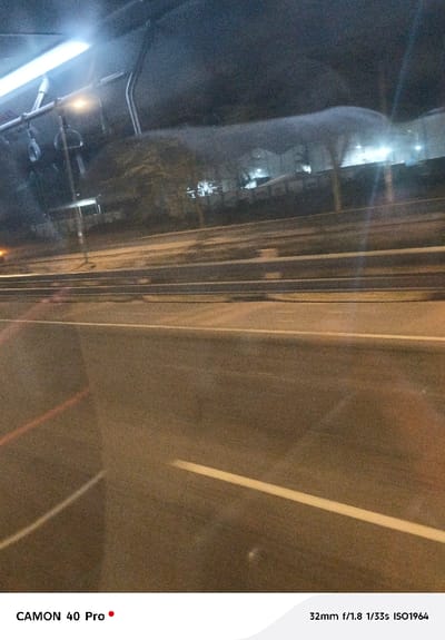 The image captures a blurry, grainy scene taken at night from inside a moving vehicle, likely a bus or car, looking out through a window. The overall impression is dark, illuminated by artificial light sources and characterized by reflections on the glass.

In the upper left quadrant, a bright, elongated light source is prominent, likely an interior light of the vehicle, casting a strong reflection. Below it, several dark, indistinct straps or handles, similar to those found in public transport, are visible, further suggesting the interior of a bus. The upper part of the image also shows dark, textured shapes that could be the vehicle's ceiling or other interior components, with subtle reflections superimposed.

The midground and background depict an outdoor scene. Multiple horizontal lines stretch across the frame, indicating a multi-lane road or highway with guardrails or barriers. A utility pole or street light pole with a faint light at its top is visible on the left side, positioned between the guardrails and the distant background. The distant background shows dark silhouettes of trees and buildings, with scattered points of artificial light emanating from windows or streetlights.

The lower half of the image is dominated by the road surface, which appears dark and somewhat wet or reflective. A distinct white horizontal line, likely a lane marker, runs across the lower-middle part of the frame. A fainter, reddish line is also barely visible near the bottom left edge. The entire outdoor scene is somewhat obscured and softened by the window's reflections and the low-light conditions, which also contribute to the image's grainy texture.

At the bottom left corner, the text "CAMON 40 Pro •" identifies the camera used. In the bottom right corner, camera settings are displayed: "32mm f/1.8 1/33s ISO1964".