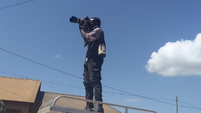 A man stands atop a silver structure, perhaps a vehicle roof rack, against a bright blue sky with a fluffy white cloud. He is taking a photograph with a professional camera. He wears a patterned shirt, dark pants with cargo pockets, and a white bag hangs at his side. Overhead wires are visible above a building with a corrugated roof.