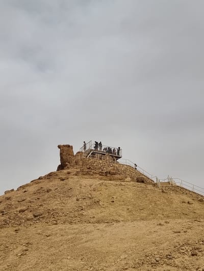 The image depicts a cliffside structure located in Mitzpe Ramon, Israel. Atop the cliff, a viewing platform with a white metal railing is populated by several people, estimated to be around a dozen. The cliff face is composed of layered rock and soil, showing a mix of textures. The sky is overcast and grey. The image is taken from a low angle, emphasizing the height and verticality of the cliff.