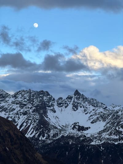 The image displays a dramatic alpine landscape, likely captured during dawn or dusk, given the presence of a bright moon in a partially blue sky. The composition is vertically oriented, with towering mountains occupying the right side of the frame and the sky filling the left.

The mountains are rugged and grand, characterized by jagged, rocky peaks heavily covered in white snow, particularly on their upper reaches. Darker, exposed rock faces are also visible, contrasting sharply with the snow. A dark, less distinct mountain slope or hillside appears in the lower right corner.

The sky on the left side of the image is a clear, light blue near the top-left, transitioning into a more overcast expanse with various cloud formations. Some clouds are brightly illuminated, appearing white or light grey, while others are darker grey, suggesting depth and varying light penetration. A prominent, nearly full moon glows brightly in the upper-left portion of the blue sky, adding to the serene yet grand atmosphere.

The lighting suggests the sun is low on the horizon, illuminating specific parts of the clouds and the snow-capped peaks, and casting shadows that define the mountainous terrain. The scene depicts a tranquil, majestic natural environment. There is no visible human activity or text. The location context provided is Poschiavo, Switzerland, which aligns with the alpine scenery.
