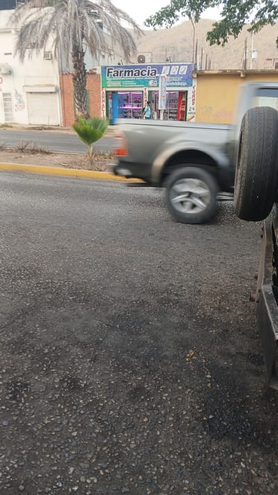 The image shows a street scene in Puerto La Cruz, Venezuela. A vehicle, partially visible, occupies the upper section of the frame. In the background, a pharmacy named "Farmacia" is present, identified by its signage and storefront. The street appears to be paved and is bordered by a yellow curb. A palm tree and other vegetation are visible along the roadside. A building is on the opposite side of the street. In the background, a mountain can be seen.