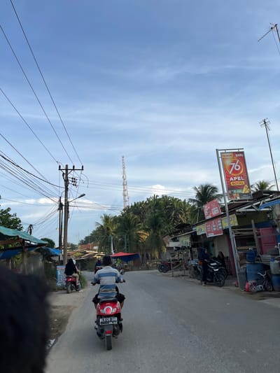 In Alurcucur, Indonesia, a person rides a red scooter down a paved road. The scooter is a red Honda Beat with a license plate reading BL 5129 UAB, dated 08-22. The rider wears a striped blue and white shirt and black shorts. To the right of the road, there is a building with a tall, red and white banner advertising "DIARUM BARU 76 APEL ROYAL". Several other signs and structures line the road, including a utility pole with numerous wires and a distant cell tower. Tropical trees, including palm trees, are visible in the background. Other people and scooters are present on the side of the road and further down the street. The sky is partly cloudy with a bright blue hue.