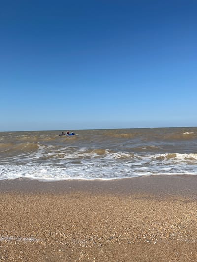 The image depicts a serene beach scene in Dolzhanskaya, Russia. In the foreground, a sandy beach is visible, with the gentle waves of the sea lapping at the shore. The water is a murky brown, indicating sediment stirred up from the seabed. In the mid-ground, a small inflatable boat with three individuals is seen navigating the choppy waters. The individuals appear to be enjoying a recreational activity, possibly tubing or simply cruising. The sky above is a clear, vibrant blue, suggesting a pleasant day for outdoor activities. The overall atmosphere of the image is one of tranquility and leisure, capturing a typical day at the beach in this coastal town.