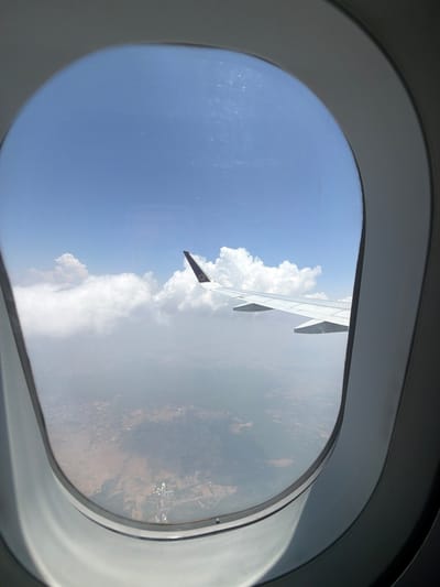 The image is taken from an airplane window, looking out at the sky and the landscape below. A portion of the airplane's wing is visible on the right side of the frame. The wing is white with a purple logo on its tip. The sky is blue with scattered white clouds. Below the clouds, a hazy landscape of a city or town is visible, with buildings and roads. Based on the purple logo on the wing tip, this appears to be an IndiGo Airlines plane. IndiGo Airlines is a major airline in India, and the landscape below suggests a view over a populated area. Without further information or a specific landmark, pinpointing the exact location within Gurgaon, India is not possible solely from this image. However, given the urban landscape and the presence of an Indian airline, it is plausible that the image was taken while flying over or near Gurgaon, a major city in the National Capital Region of India.
