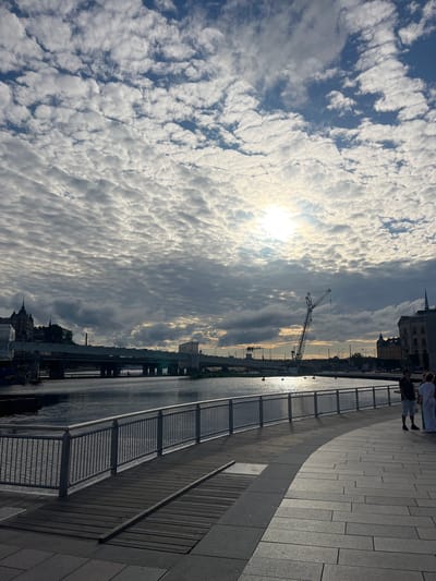 This media file, taken in Stockholm, Sweden, depicts a waterfront scene with a bright sky filled with textured clouds. The sun is visible, suggesting it is either sunrise or sunset. A paved walkway borders the water, separated by a metal railing. A few people are walking along the walkway, their forms silhouetted against the light. In the distance, across the water, buildings and a construction crane are visible, indicating ongoing development. The water reflects the sunlight, creating a shimmering effect. The overall mood is serene, with the dynamic cloudscape and the calm water contributing to a sense of peaceful observation. No visible text is present in the image.