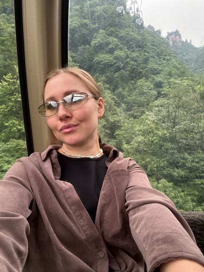 A young woman wearing glasses, a black top, a beaded necklace, and a brown corduroy jacket is shown in a medium shot inside a cable car. The background is a dense, green forest with other cable cars visible. The location is in Wulingyuan, China. The woman is the primary subject and is the focus of the image.